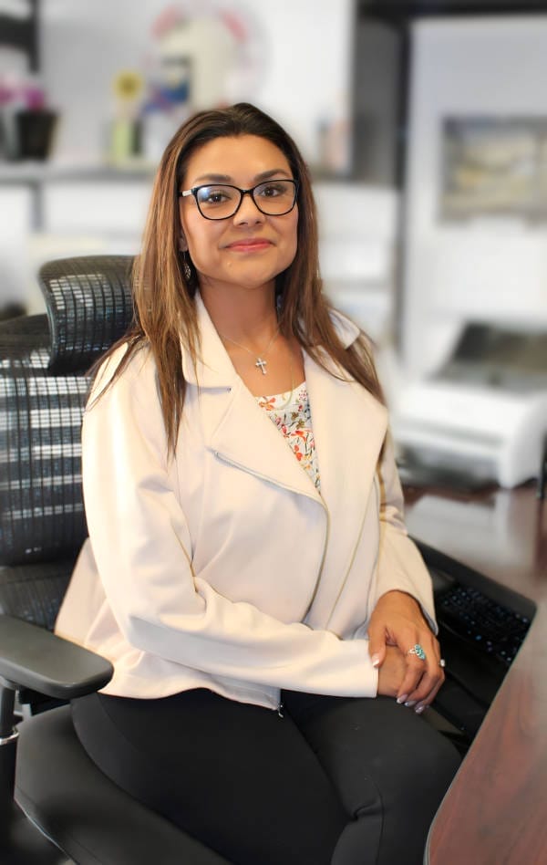 A professional woman sitting at her desk in an office environment, wearing glasses and a stylish jacket.